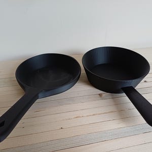 May include: Two black frying pans with long handles. One pan is round with a flat bottom, and the other is oval with a textured interior. Both pans are on a light wood surface.