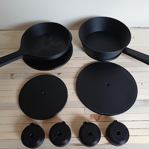 May include: A black cookware set comprising two pans with handles, two circular lids, and four small, round components. The items are arranged on a light-coloured wooden surface. The pans have a matte finish.
