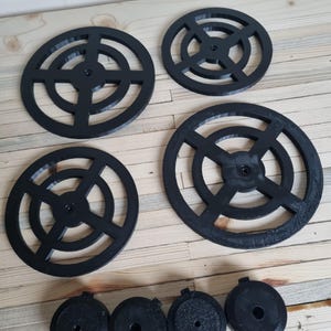 May include: Four black, circular coasters with a geometric design, and four smaller black, circular objects. The coasters have a central hole and concentric rings and crossbars. The items are arranged on a light wood surface.