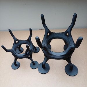 May include: Two black, 3D-printed plant stands. The stands have a unique, open design with curved legs and circular bases. The larger stand is approximately 15 cm tall, while the smaller one is about 10 cm tall. They are designed to hold plants.