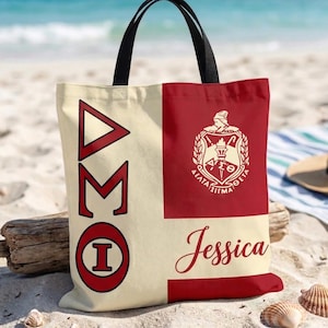 May include: A canvas tote bag with black handles, resting on a sandy beach. The bag is divided into two sections: one cream-colored with red Greek letters and a circle, and the other red with a white emblem and the name "Jessica".