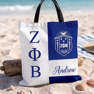 May include: A white and blue tote bag with black handles, featuring the Greek letters ZΦB in blue on one side and a crest with text on the other. The name "Andrew" is printed in script. The bag is on a sandy beach.
