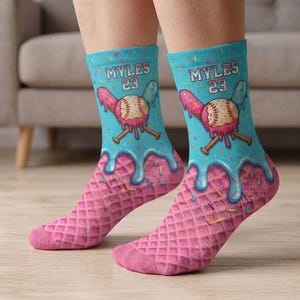 Op de afbeelding: Een paar novelty sokken met een honkbalthema. De sokken hebben een lichtblauwe achtergrond met de naam "MYLES" en het nummer "23". Een honkbal met gekruiste knuppels en een druipend ijsontwerp is op de sokken gedrukt. Het onderste deel van de sokken is roze met een wafelpatroon.