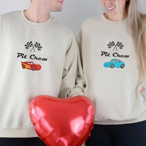 May include: Two beige sweatshirts with the text "Pit Crew" and a checkered flag design. One sweatshirt features a red car graphic, the other a blue car. A red heart-shaped balloon is held in front.