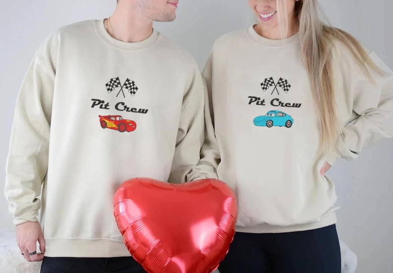 May include: Two beige sweatshirts with the text "Pit Crew" and a checkered flag design. One sweatshirt features a red car graphic, the other a blue car. A red heart-shaped balloon is held in front.