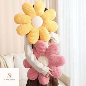 Op de afbeelding: Twee grote bloemvormige kussens. Eén is geel met een wit centrum, de andere is roze met een wit centrum. De kussens hebben een getextureerde, pluche uitstraling. De kussens worden door een persoon vastgehouden.