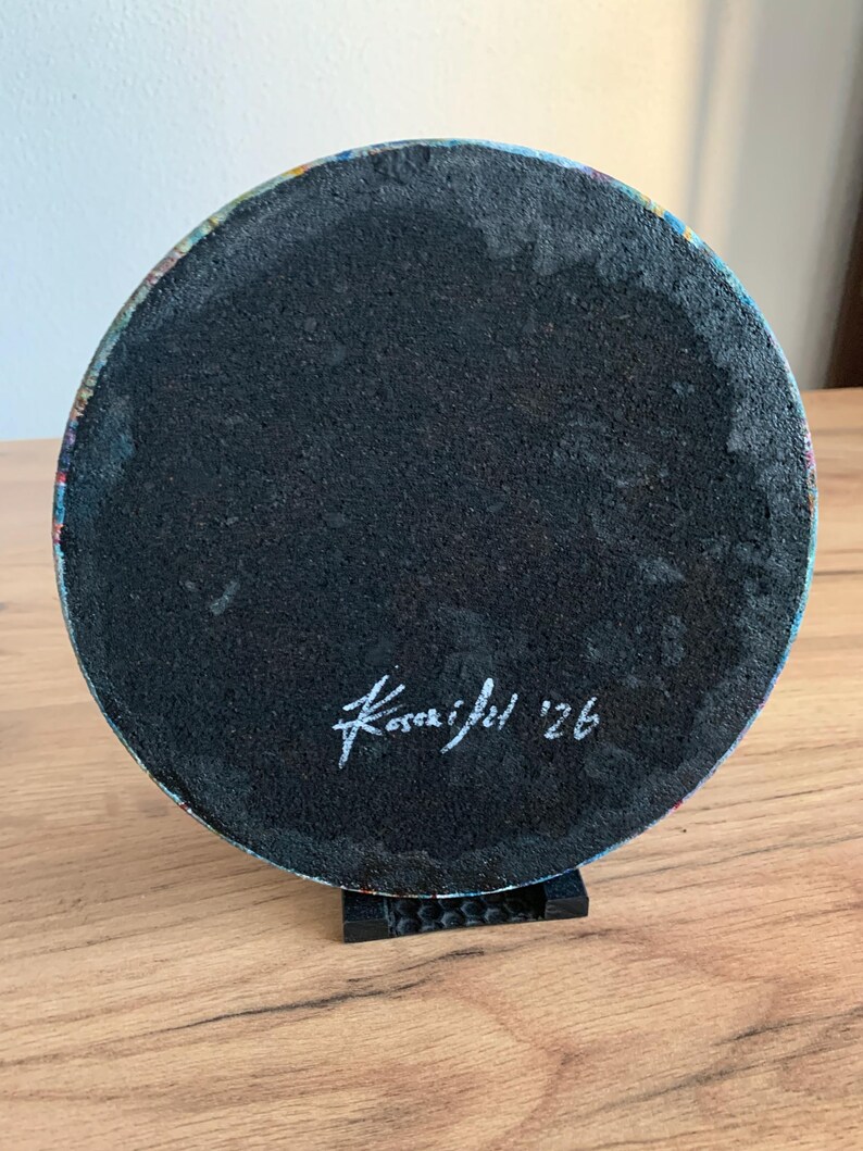 May include: A round, black object with a textured surface, possibly a coaster or artwork base. The edge shows a colorful, abstract design. The object is supported by a small, black stand. The artist's signature and the year '26 are visible.