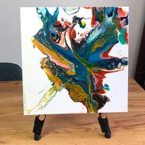 Könnte beinhalten: Abstrakte Acryl-Gießmalerei auf einer weißen Leinwand, präsentiert auf einer kleinen schwarzen Staffelei. Das Kunstwerk zeigt eine lebendige Mischung aus Farben, darunter Blau, Grün, Rot und Gelb, die ein dynamisches und fließendes Design erzeugt. Die Leinwand ist etwa 30 cm groß.
