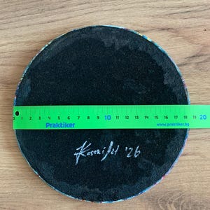 May include: A round, black coaster with a colorful patterned edge. A green ruler shows the coaster is approximately 8 inches in diameter. The coaster has the signature "Kasai fel '26" in white.