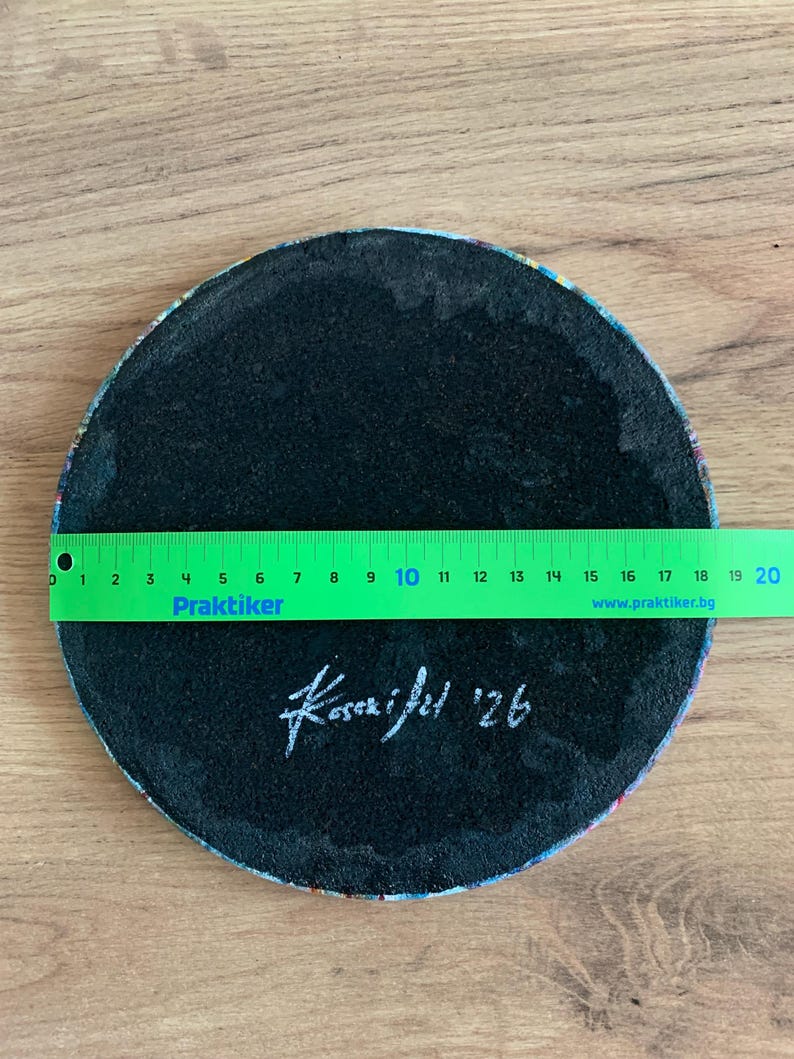 May include: A round, black coaster with a colorful patterned edge. A green ruler shows the coaster is approximately 8 inches in diameter. The coaster has the signature "Kasai fel '26" in white.