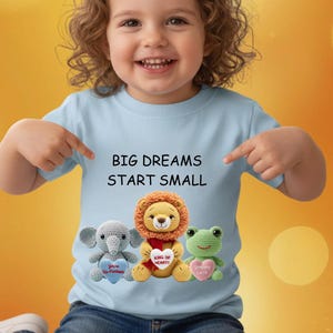 Pode incluir: Camiseta azul clara com o texto "BIG DREAMS START SMALL". A camiseta apresenta três bichos de pelúcia de crochê: um elefante, um leão e um sapo. Cada animal segura um coração.