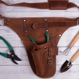 Op de afbeelding: Bruine leren tuingereedschapsriem met een riem, zakken en metalen hardware. Inclusief groene en zwarte snoeischaar, een kleine handhark en een troffel. De riem is ontworpen om om de taille te worden gedragen.