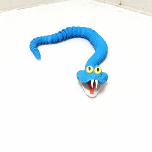 Könnte beinhalten: Eine blaue Spielzeugschlange mit einem segmentierten Körper und einem cartoonartigen Kopf. Der Schlangenkopf hat gelbe Augen, weiße Zähne und eine rote Zunge. Das Spielzeug besteht aus einem flexiblen Material.