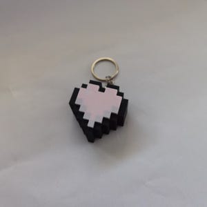 Könnte beinhalten: Ein pixelartiger herzförmiger Schlüsselanhänger. Das Herz ist schwarz mit einem hellrosa Zentrum und weißen Akzenten. Es ist an einem goldfarbenen Schlüsselring befestigt. Das Design erinnert an Retro-Videospielgrafiken.