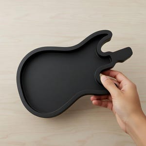 Puede incluir: Una bandeja negra con forma de guitarra y superficie lisa. La bandeja es sostenida por una mano, mostrando su diseño. La bandeja está hecha de un material sólido y está diseñada para guardar objetos pequeños.