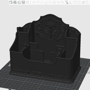 May include: A matte black desktop organizer featuring a central shield-shaped back panel embossed with a detailed rose motif. The structure includes multiple compartments of varying heights for storage, with additional rose vine relief detailing.