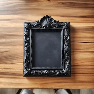 May include: A black, ornate picture frame with a rectangular opening. The frame has intricate floral and scrollwork designs, and a shell-shaped crest at the top. The frame is sitting on a wooden surface.