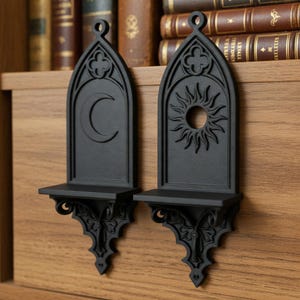 May include: Two black gothic-style wall shelves. One features a crescent moon, the other a sun design. Each shelf has a small ledge and ornate detailing. The shelves are mounted on a wooden surface, with books in the background.