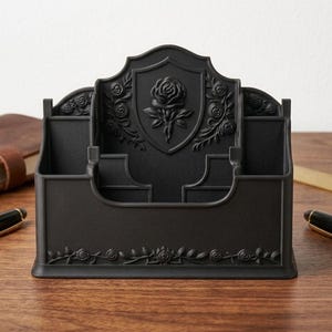 May include: A matte black desk organizer featuring a central shield-shaped panel embossed with a rose motif. The organizer has multiple compartments for stationery and is decorated with additional rose and vine relief patterns along the front base.