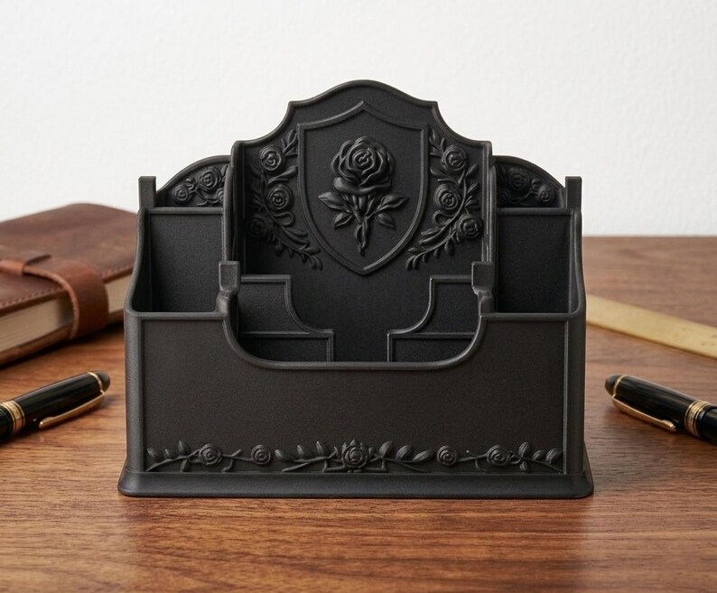 May include: A matte black desk organizer featuring a central shield-shaped panel embossed with a rose motif. The organizer has multiple compartments for stationery and is decorated with additional rose and vine relief patterns along the front base.