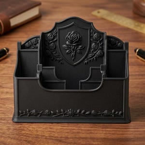 May include: A matte black desk organizer featuring a central shield emblem with a raised rose relief. The organizer has multiple compartments for stationery and is decorated with intricate rose vine patterns along the base and sides.