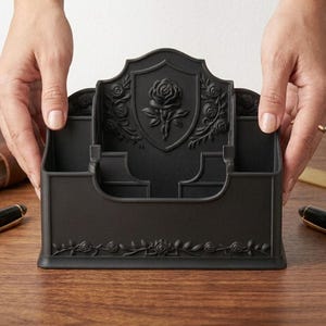 May include: A matte black desk organizer featuring a central shield motif embossed with a rose. The organizer includes multiple compartments for stationery and is decorated with intricate rose vine patterns along the front base and side panels.