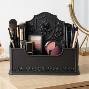 May include: A matte black plastic makeup organizer featuring a raised rose emblem on the back panel and floral vine detailing along the base. The organizer contains various compartments holding makeup brushes, eyeshadow palettes, lipstick, and sponges.