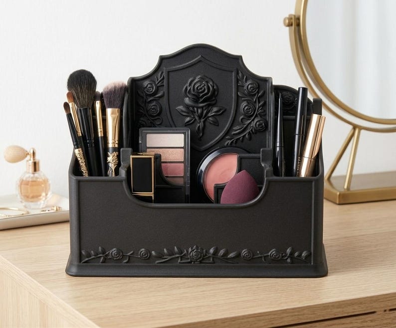May include: A matte black plastic makeup organizer featuring a raised rose emblem on the back panel and floral vine detailing along the base. The organizer contains various compartments holding makeup brushes, eyeshadow palettes, lipstick, and sponges.