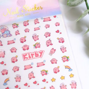 K&ouml;nnte beinhalten: Nagelkunst-Aufkleberbogen mit rosa Kirby-Charakteren, Sternen, Herzen und Regenbogen. Das Blatt enth&auml;lt das Wort "Kirby" in Rosa und den Text "Nail Sticker" in Gold.