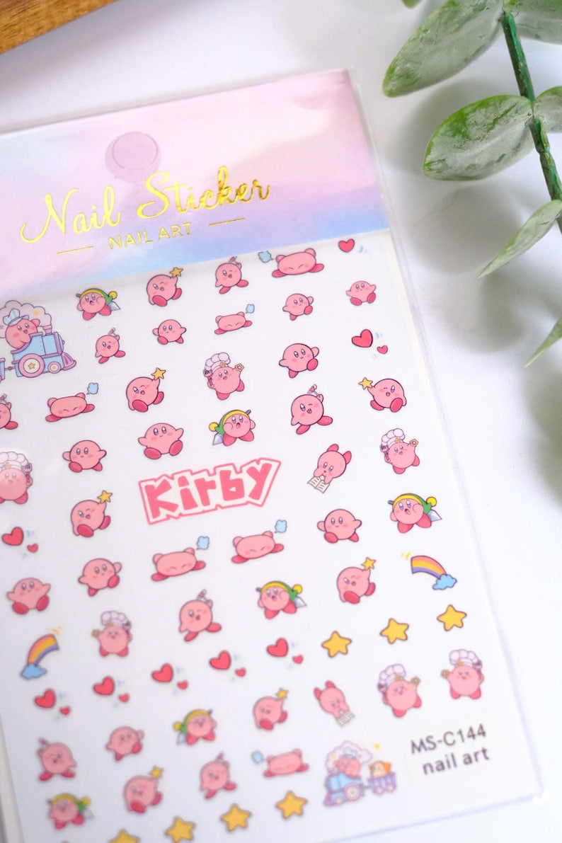 K&ouml;nnte beinhalten: Nagelkunst-Aufkleberbogen mit rosa Kirby-Charakteren, Sternen, Herzen und Regenbogen. Das Blatt enth&auml;lt das Wort "Kirby" in Rosa und den Text "Nail Sticker" in Gold.