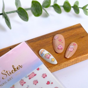 K&ouml;nnte beinhalten: Nagelkunst-Aufkleber und bemalte N&auml;gel mit einer rosa Cartoonfigur. Die Aufkleber befinden sich auf einem Bogen mit pastellfarbenem Hintergrund. Drei N&auml;gel sind mit der Figur und anderen Designs bemalt. Ein Zweig mit gr&uuml;nen Bl&auml;ttern befindet sich im Hintergrund.