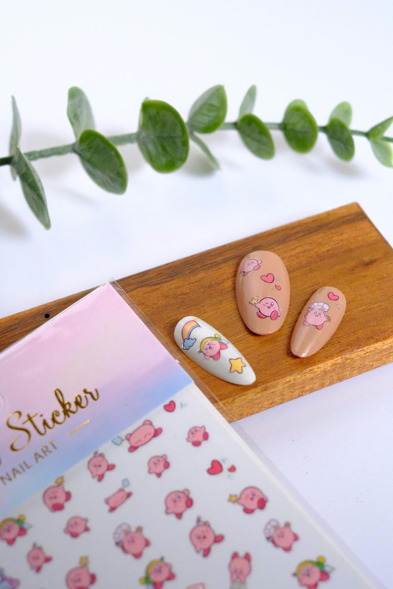K&ouml;nnte beinhalten: Nagelkunst-Aufkleber und bemalte N&auml;gel mit einer rosa Cartoonfigur. Die Aufkleber befinden sich auf einem Bogen mit pastellfarbenem Hintergrund. Drei N&auml;gel sind mit der Figur und anderen Designs bemalt. Ein Zweig mit gr&uuml;nen Bl&auml;ttern befindet sich im Hintergrund.