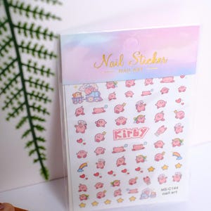 K&ouml;nnte beinhalten: Eine Packung Nagelkunst-Aufkleber mit der Figur Kirby. Die Aufkleber zeigen verschiedene rosa Kirby-Designs, Herzen und Sterne. Die Verpackung tr&auml;gt die Worte "Nail Sticker" in goldener Schrift.
