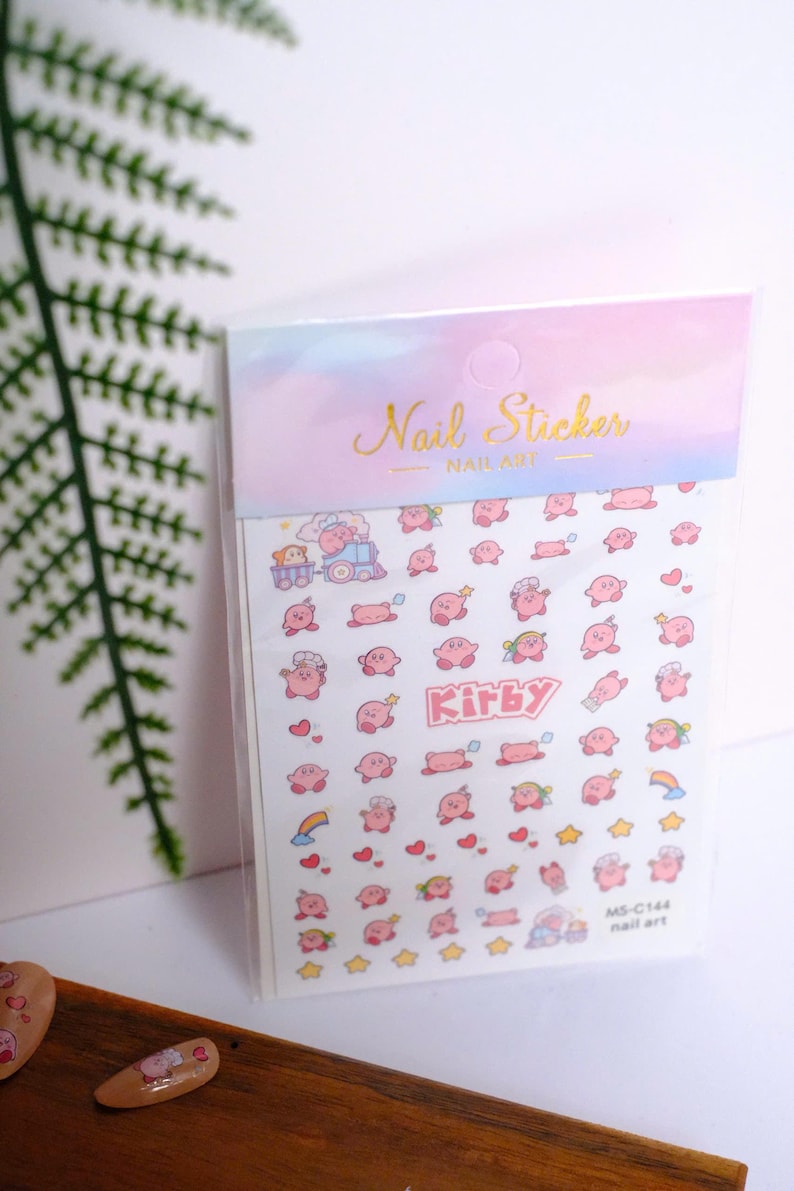 K&ouml;nnte beinhalten: Eine Packung Nagelkunst-Aufkleber mit der Figur Kirby. Die Aufkleber zeigen verschiedene rosa Kirby-Designs, Herzen und Sterne. Die Verpackung tr&auml;gt die Worte "Nail Sticker" in goldener Schrift.