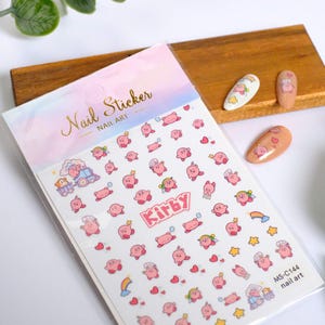 K&ouml;nnte beinhalten: Nagelkunst-Aufkleber mit rosa Kirby-Charakteren in verschiedenen Posen, mit Sternen, Herzen und Regenbogen. Auf der Verpackung steht "Nail Sticker Nail Art" und der Text "Kirby". Die Aufkleber befinden sich auf wei&szlig;em Hintergrund.