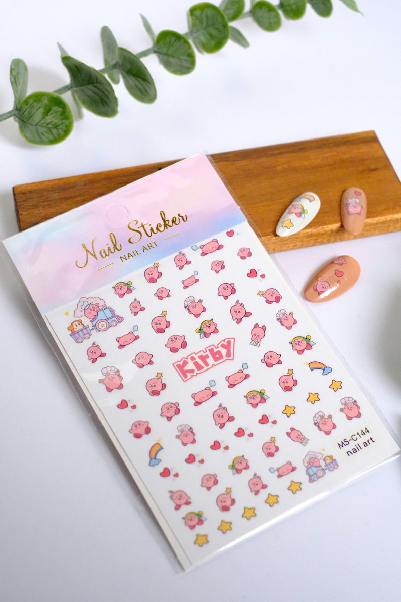 K&ouml;nnte beinhalten: Nagelkunst-Aufkleber mit rosa Kirby-Charakteren in verschiedenen Posen, mit Sternen, Herzen und Regenbogen. Auf der Verpackung steht "Nail Sticker Nail Art" und der Text "Kirby". Die Aufkleber befinden sich auf wei&szlig;em Hintergrund.