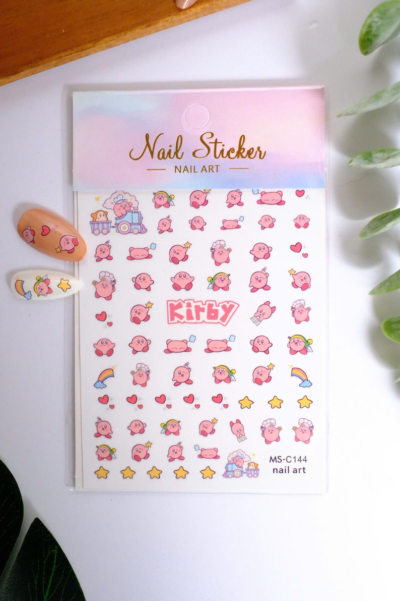 K&ouml;nnte beinhalten: Nagelkunst-Aufkleber mit einem rosa Cartoon-Charakter in verschiedenen Posen, Herzen, Sternen und Regenbogen. Das Blatt enth&auml;lt den Text "Kirby" und "Nail Sticker - Nail Art". Zwei Nagelkunst-Beispiele sind links abgebildet.