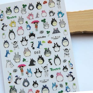 Può includere: Foglio di adesivi con vari personaggi di Totoro in diverse pose e colori. Gli adesivi includono immagini di Totoro, una ragazza con un ombrello, funghi e altri elementi correlati. Il testo "Totoro" e il numero "062" sono anch'essi visibili.