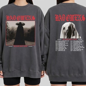 Può includere: Felpa grigio scuro con il nome della band "BAD OMENS" in lettere gotiche rosse. La parte anteriore mostra l'immagine di una figura con mantello e cappello neri. Il retro presenta una figura spettrale e un programma del tour.