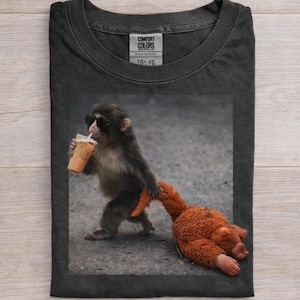 May include: Dark gray t-shirt featuring a photo of a monkey wearing sunglasses and holding a beverage. The monkey is also dragging a stuffed animal. The shirt has a Comfort Colors tag.