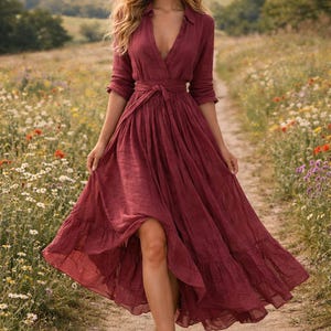 May include: A burgundy maxi dress with a V-neck and long sleeves. The dress has a tie-waist and a flowing skirt. The person is walking barefoot on a dirt path through a field of wildflowers.