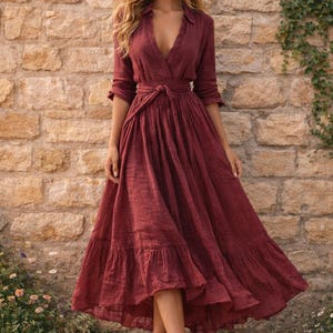 May include: A full-length burgundy dress with a V-neck and tie-waist. The dress features long sleeves and a tiered skirt, creating a bohemian style. The model is standing barefoot in front of a stone wall.