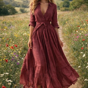 May include: A full-length burgundy dress with a V-neck and a tied waist. The dress features long sleeves and a ruffled hem. The person is walking on a dirt path through a field of wildflowers.