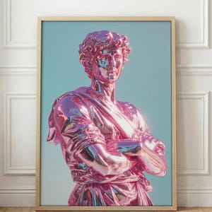 May include: A framed print featuring a metallic pink statue of a classical figure with arms crossed. The statue is set against a light blue background. The frame is a light wood color, and the artwork is displayed on a white wall and wooden floor.