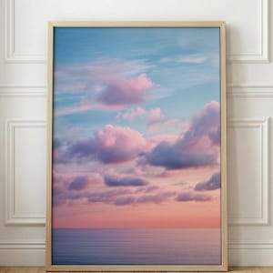 May include: A framed print featuring a serene seascape. The artwork showcases a gradient sky with hues of blue, pink, and orange, complemented by fluffy pink clouds. The ocean is a calm, muted blue. The frame is a light wood color.