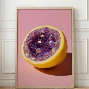 May include: A framed print featuring a halved lemon with a vibrant purple amethyst geode center. The lemon is yellow, set against a pink background. The artwork is displayed in a light-colored wooden frame, creating a striking contrast.