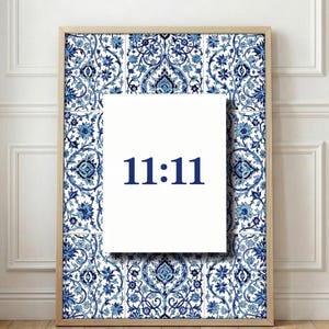 May include: A framed print featuring a white rectangle with the navy blue text "11:11" centered on a blue and white floral patterned background. The frame is a light wood color, and the print is displayed on a wooden floor.