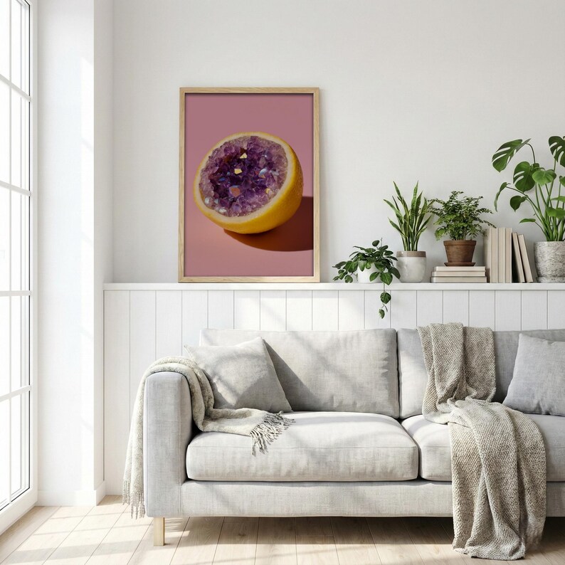 May include: A framed print featuring a halved lemon with a purple crystal geode center against a pink background. The artwork is displayed above a gray sofa with a neutral-toned throw blanket.