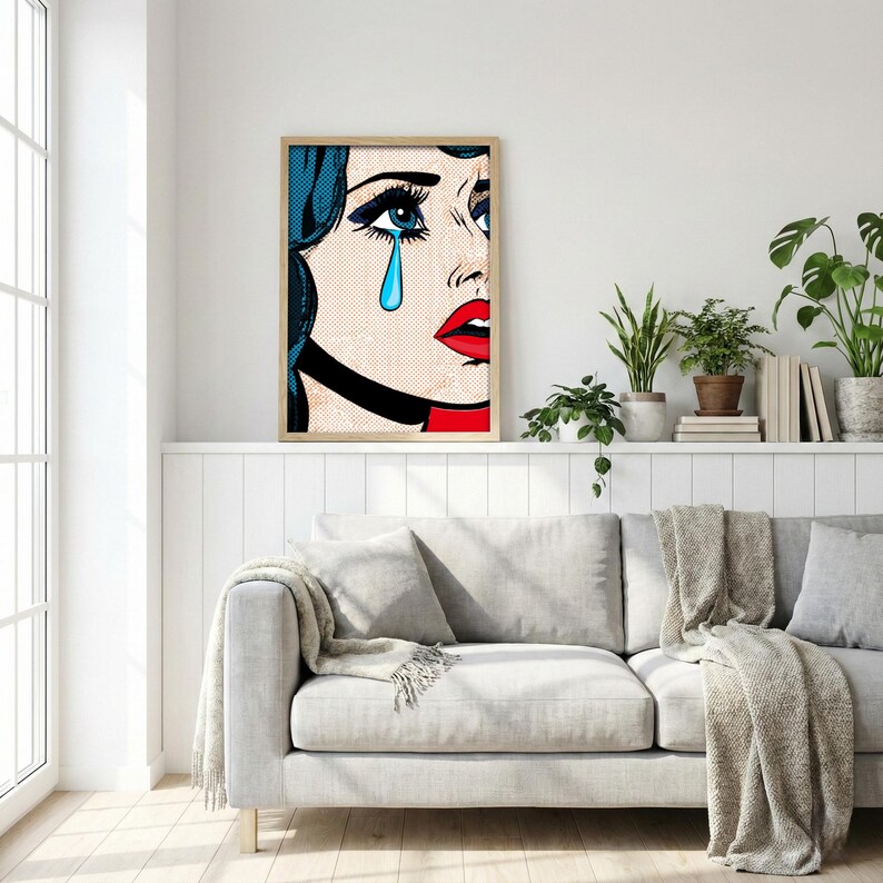 May include: Pop art print featuring a crying woman with blue hair, a single blue teardrop, and red lips. The artwork is framed in a light wood frame and hangs above a gray sofa. The style is reminiscent of comic book art.