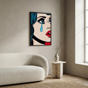May include: Pop art print of a crying woman with blue hair, a black choker, and red lips. The artwork features bold lines and a dot pattern, framed in black. The woman has a single blue teardrop.